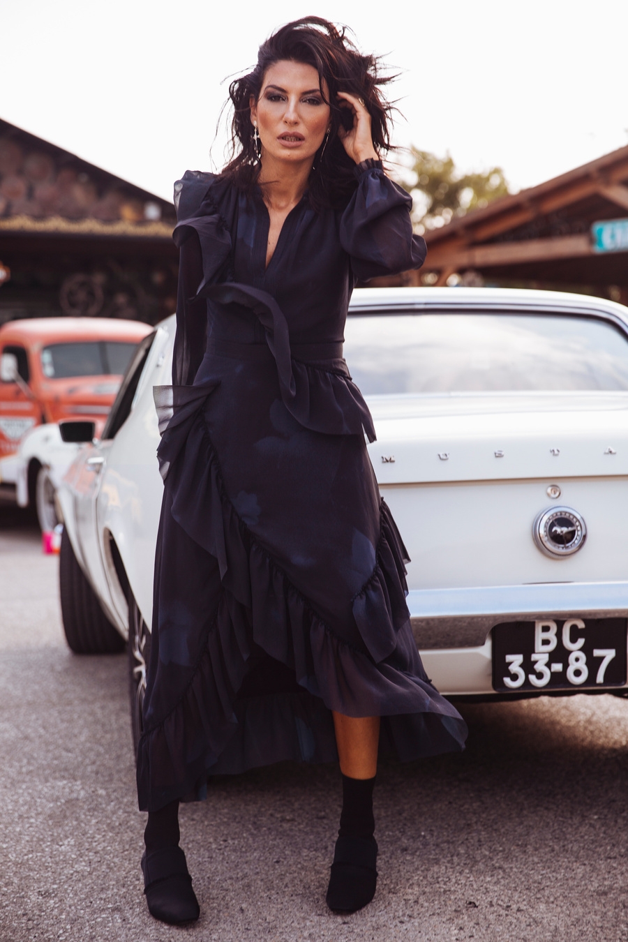 A woman wearing a long black dress and black boots stands next to an old car