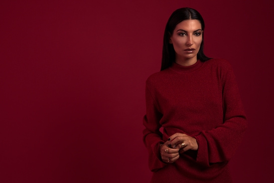 A woman standing in front of a red wall. She is wearing a red sweater, holding an object in her hand and looking into the camera