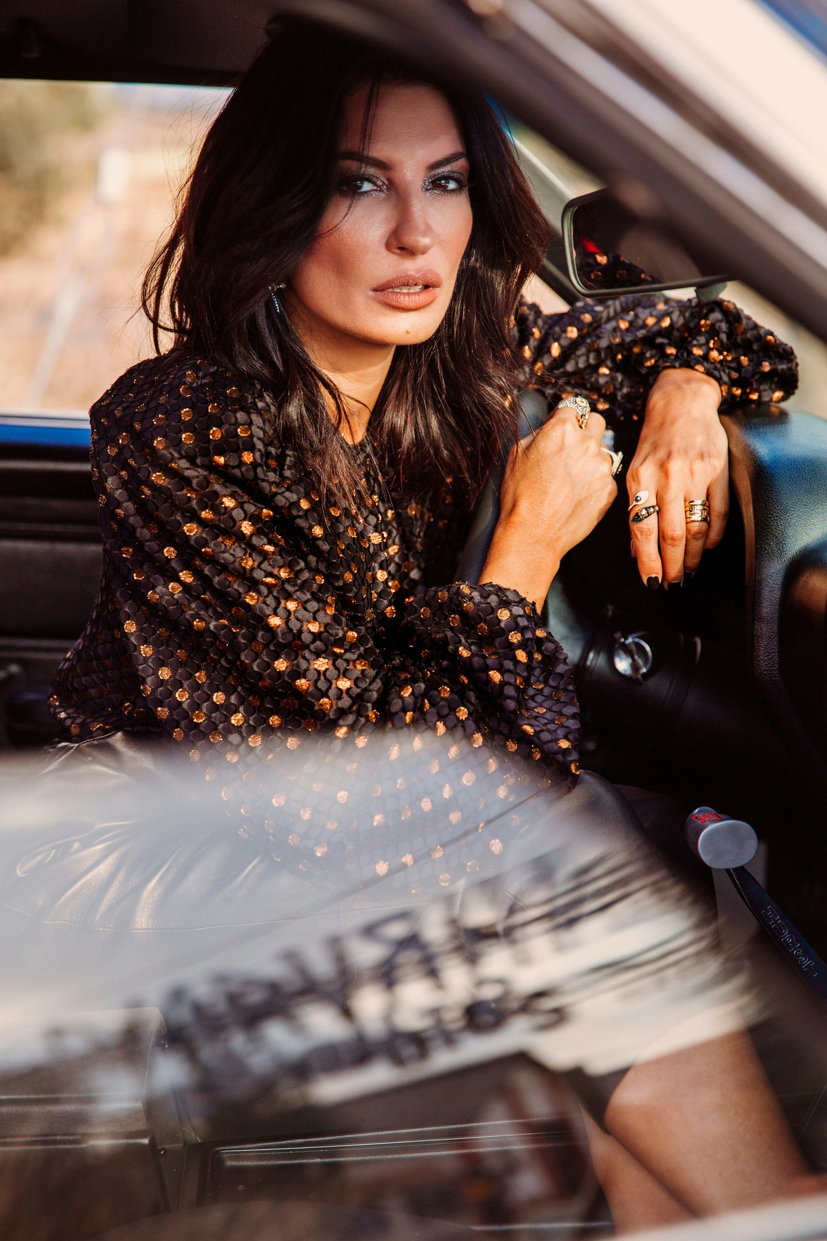 Woman leaning in her car, wearing a black top with gold rings on her fingers