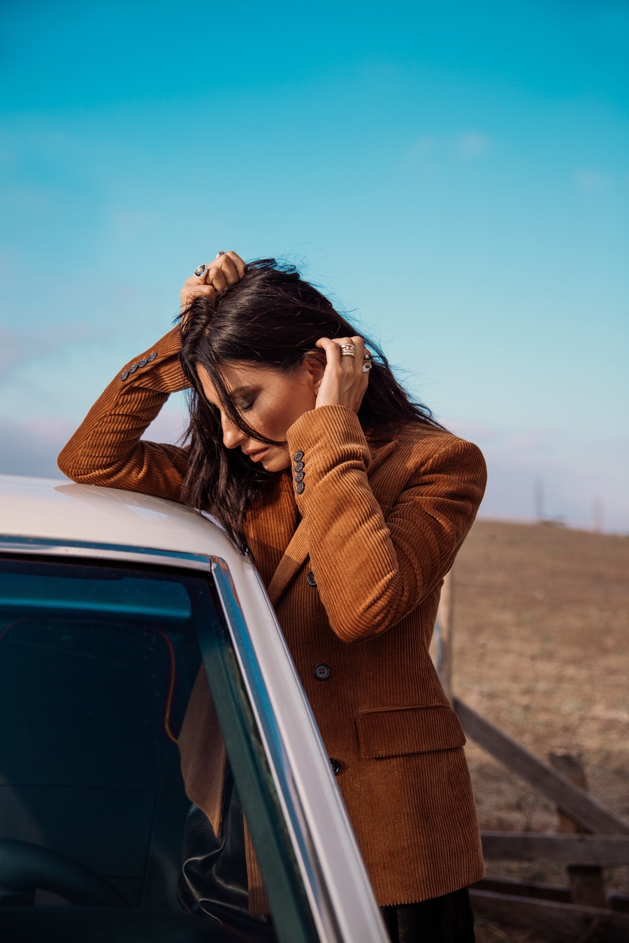 20 year old woman in a brown jacket, looking down at her car. She has long hair and is wearing black pants