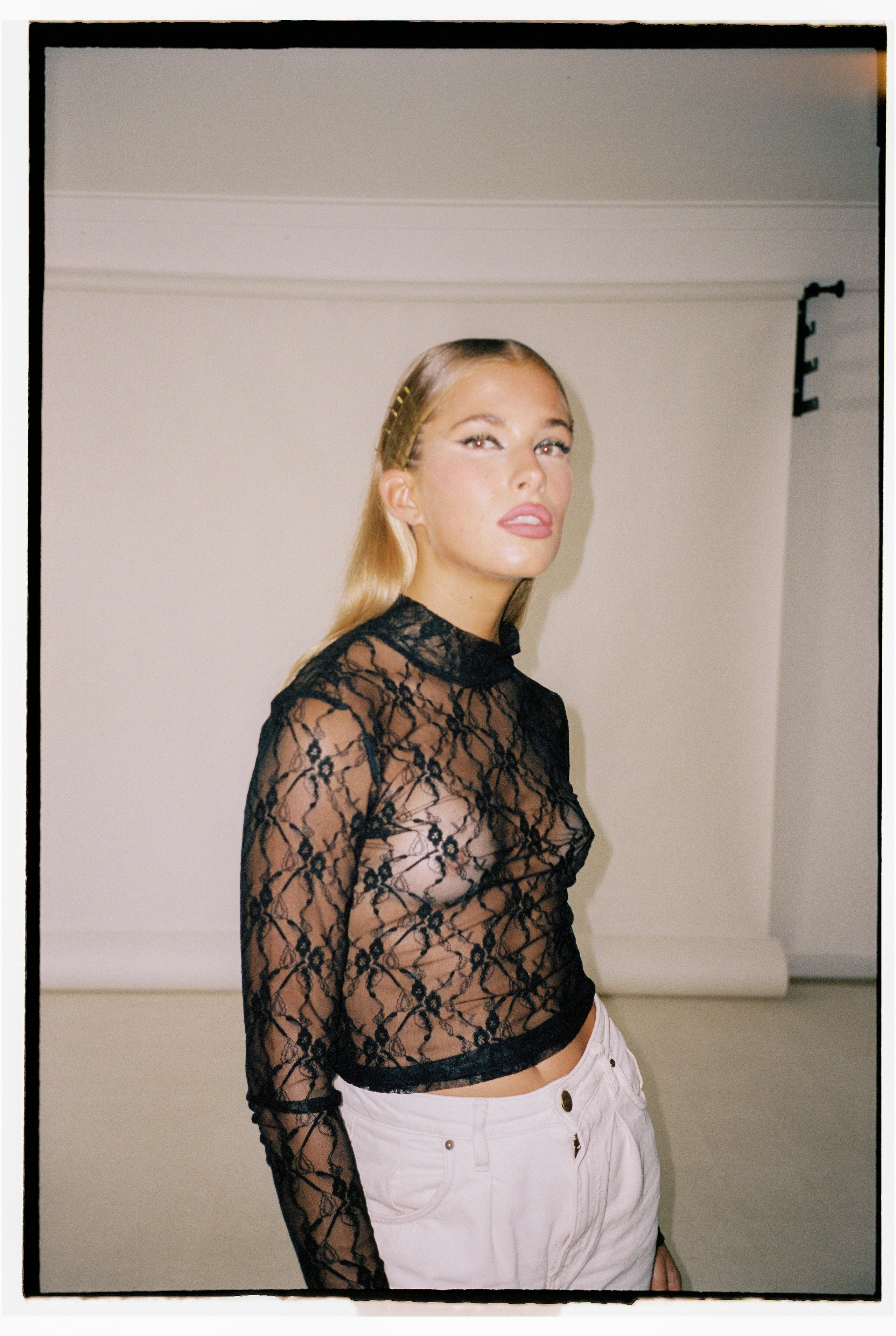 20-year old woman, wearing a black net top, looking at the camera. White walls in the background