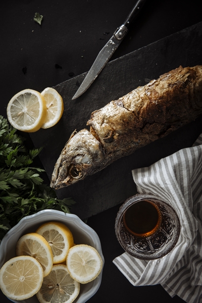 A freshly cooked fish sits on a black surface. Surrounded by lemons and herbs, the fish is ready to be served up with a side of lemonade or wine