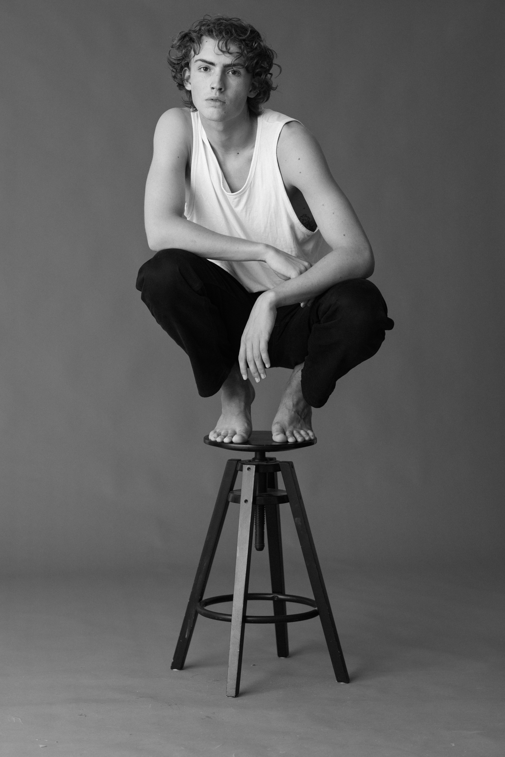 20-30 year old man sitting on stool wearing a white shirt and black pants with bare feet