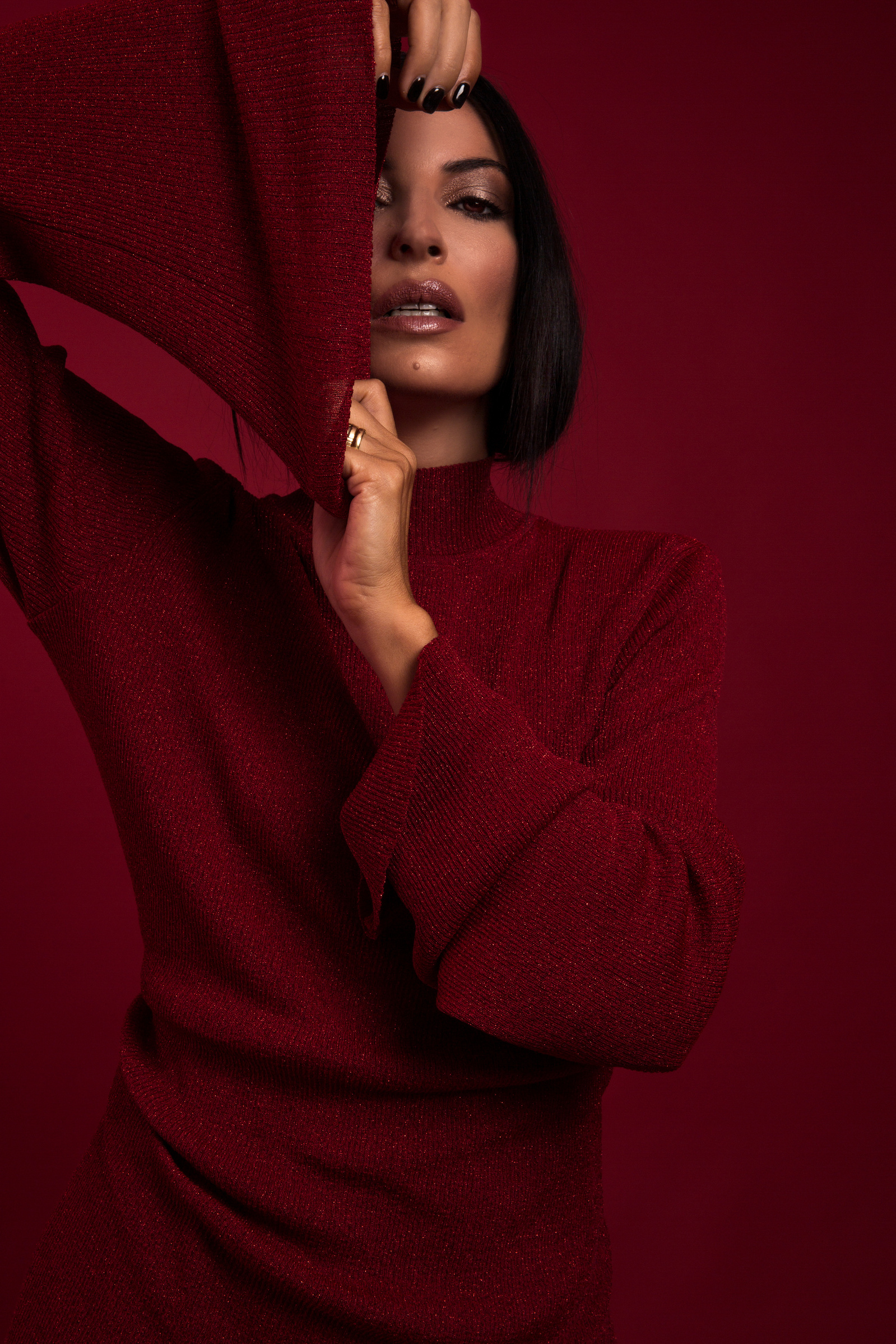 A woman wearing a red dress holds her arm up to the camera. She appears to be having a casual moment for a photo, likely in front of a red wall.