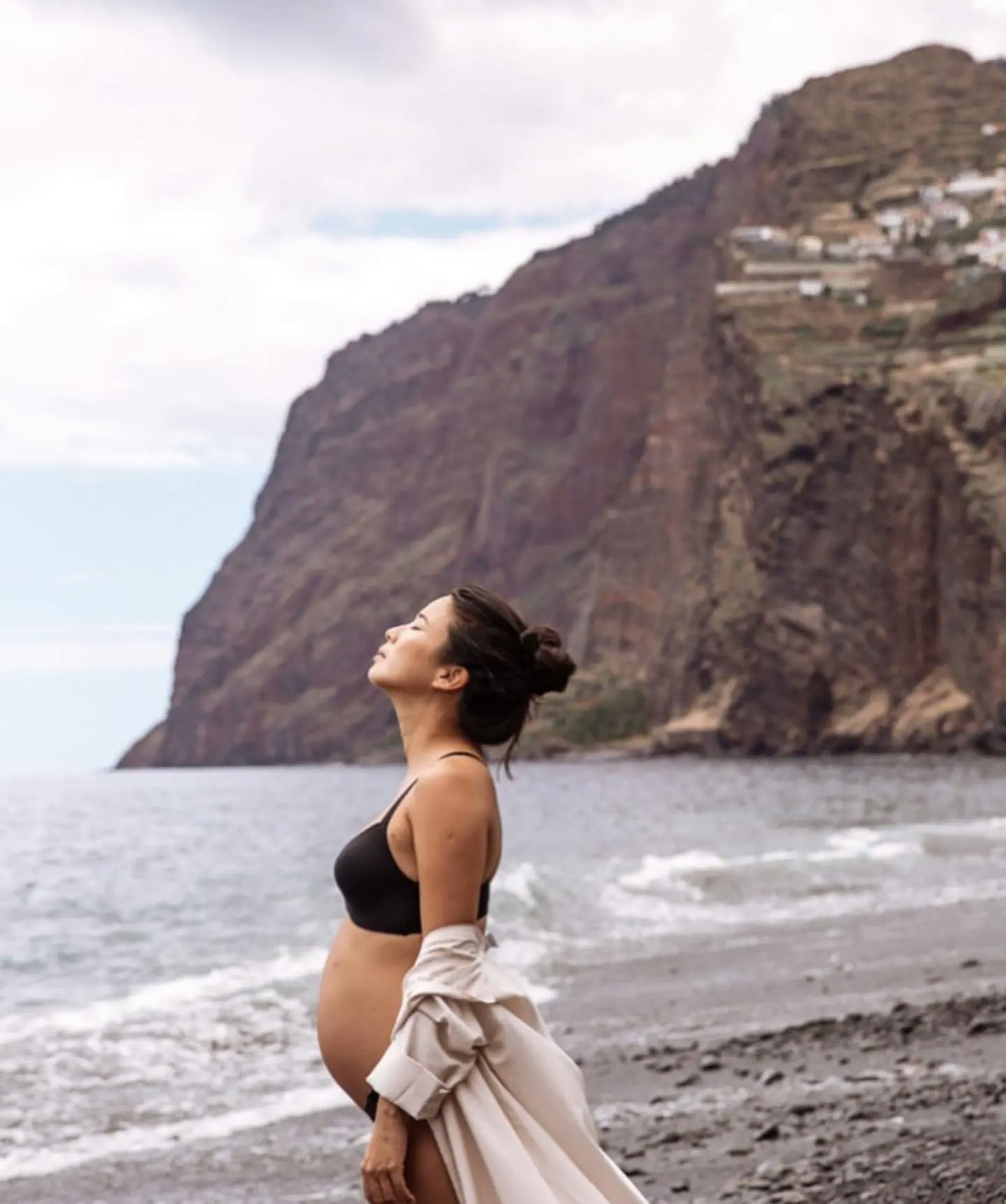 Jani Zhao with a pregnant belly, wearing a white top and black bottoms, standing on the beach near the water. Photographer: KAT V Photography.