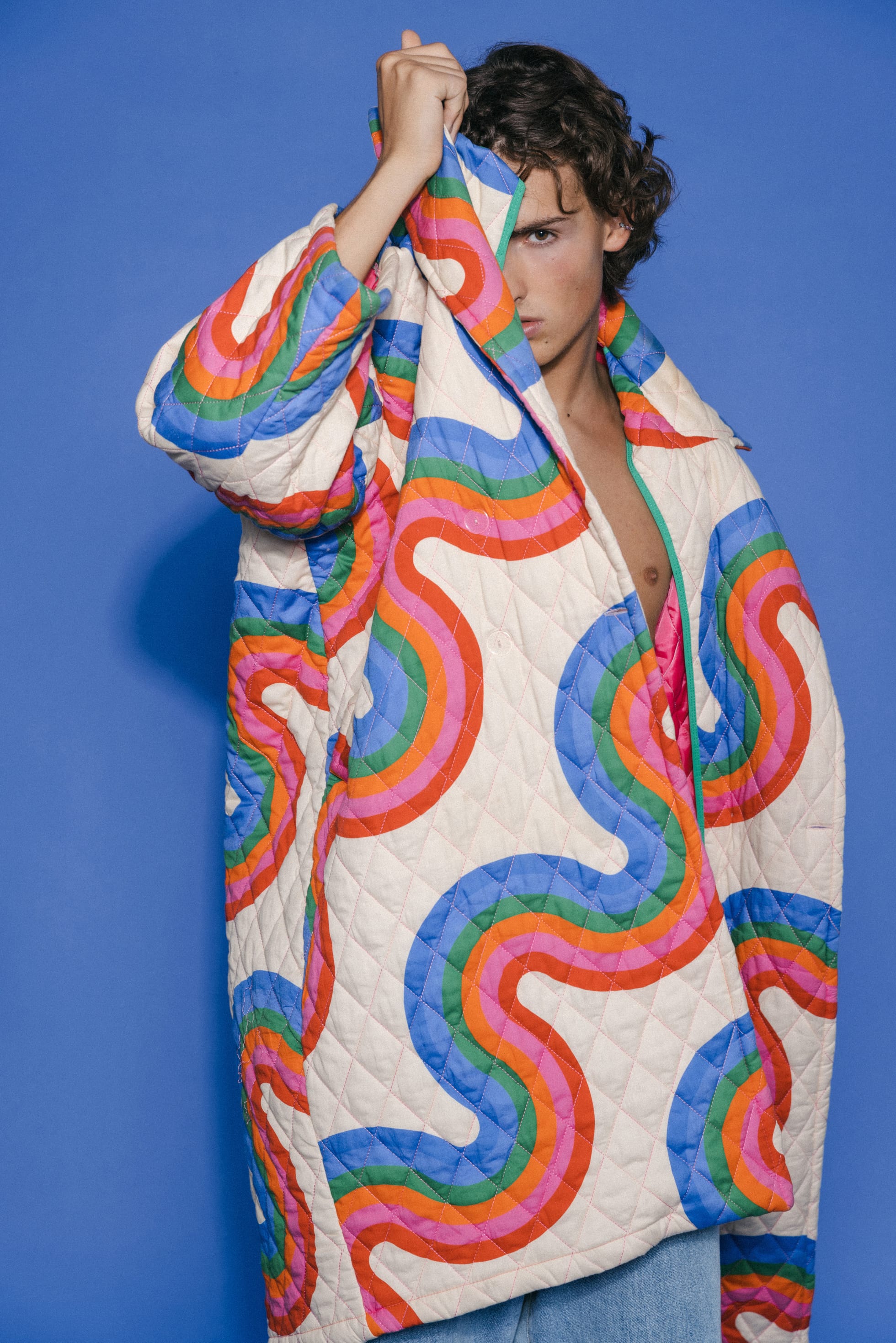 Man in open white jacket with colorful swirls posing confidently by a solid blue wall