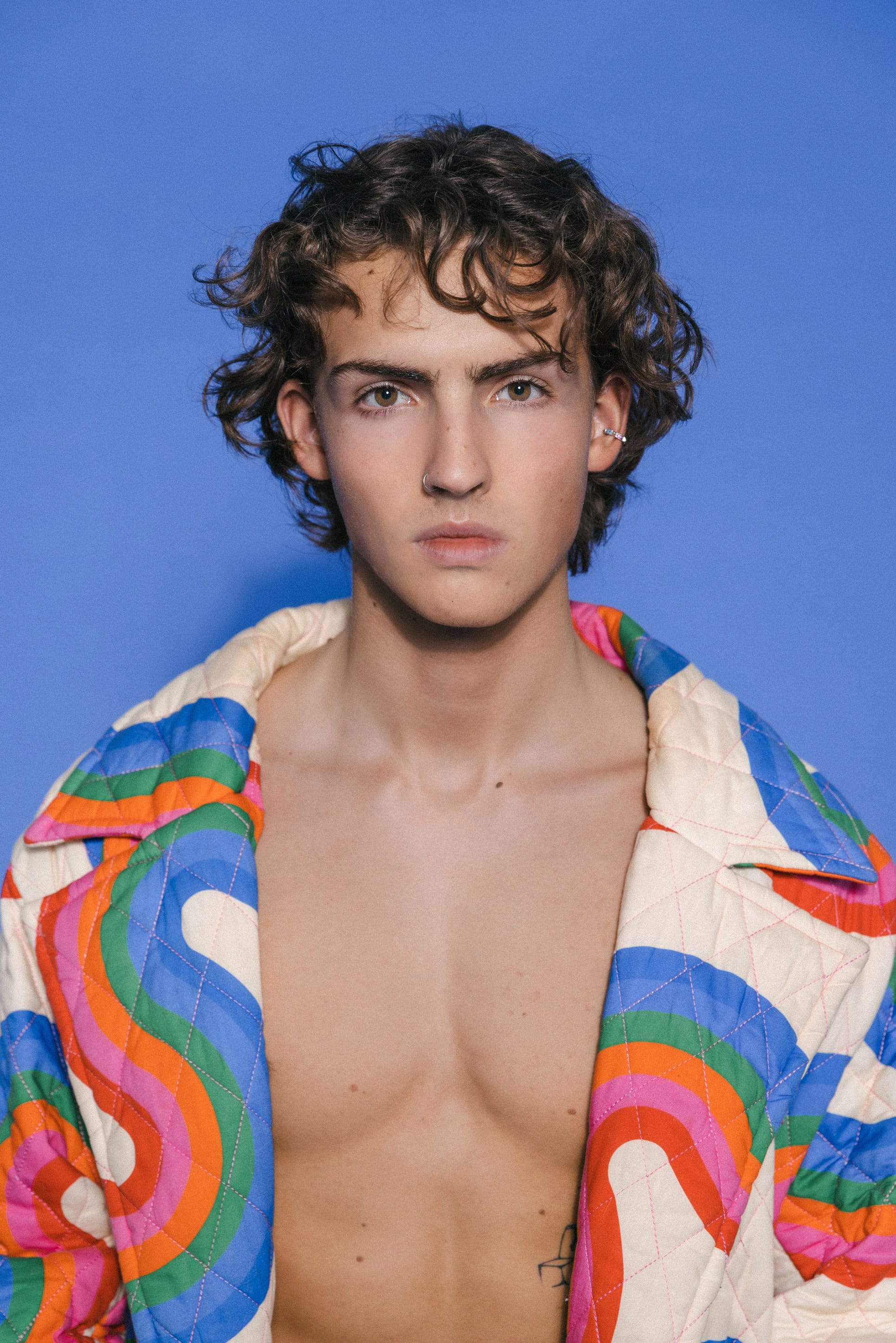 Curly-haired man in open swirly jacket posing confidently in front of a solid blue wall